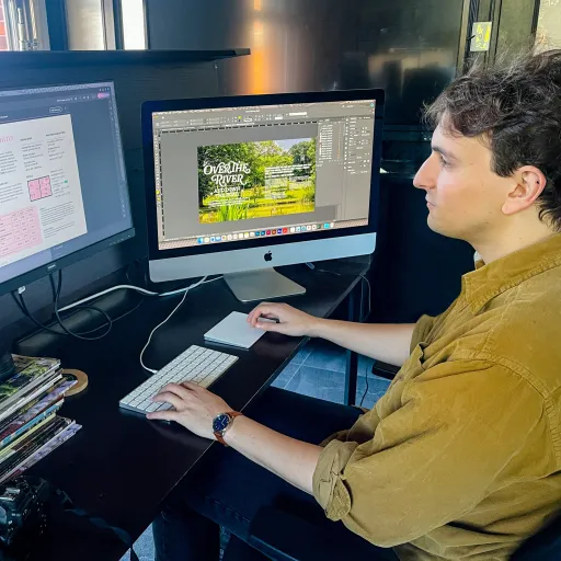 Evan Okun sits at a desk with two monitors, one showing a magazine rates page and the other an InDesign layout titled "Over the River and Down the Road."